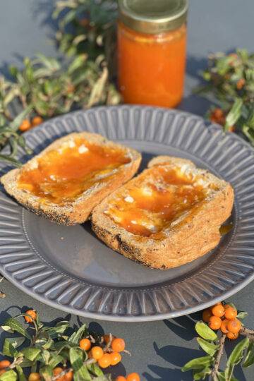 Two slices of bread with sea buckthorn jam on a dark plate, with a glass of jam and sea buckthorn branches in the background