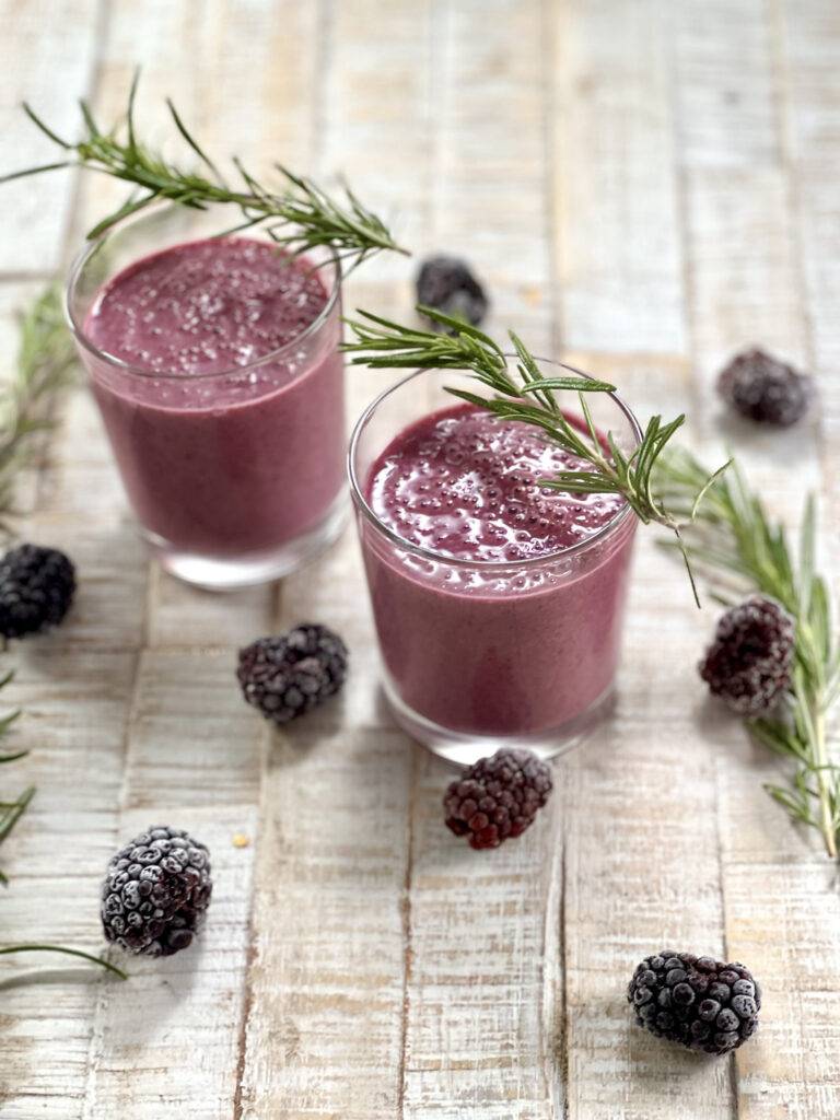 Two glasses of blackberry smoothie garnished with rosemary, and blackberries tossed around it