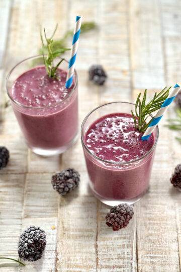 Two glasses of blackberry smoothie beautifully garnished with rosemary, blackberries and a blue straw.