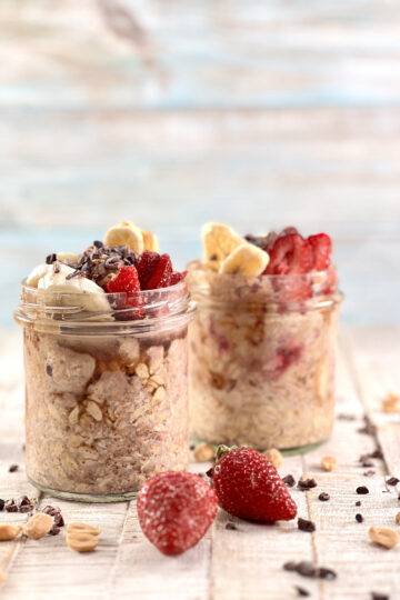 Two Jars of Vegan Peanut Butter Overnight Oats beautifully garnished, with peanuts, cacao nibs and strawberries tossed around them
