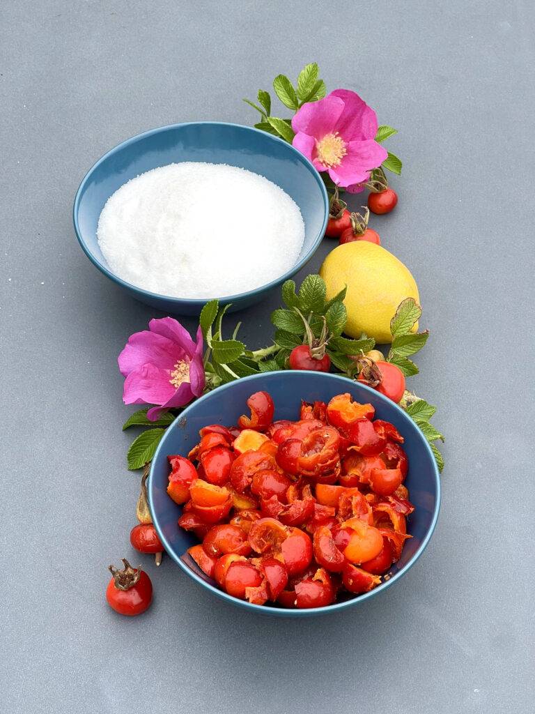 The ingredients for rose hip jam in beautiful grey plates, on a grey surface, garnished with rose leaves and flowers