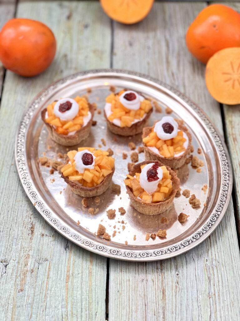 A birdseye ange of vegan persimmon tarts on a wooden underground, garnished with some vegan whipped cream and a cranberry 