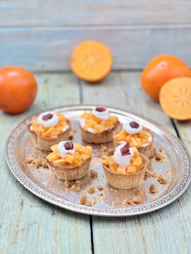 Five vegan persimmon tarts on a silver plate with sliced persimmons around them
