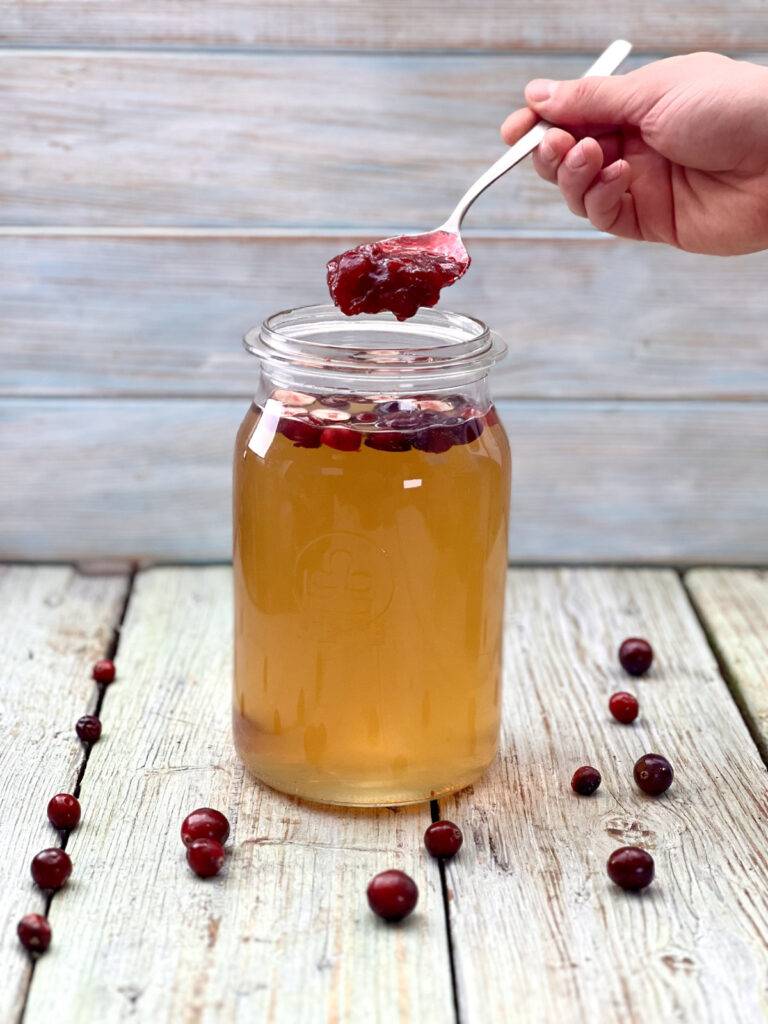 A spoonful of cranberry jam is being dropped into the fermentation jar with kombucha