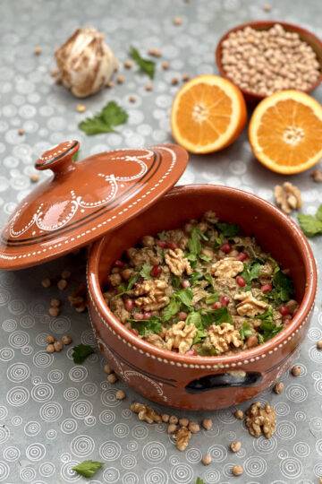 A deep clay pot with beautiful patterns full of vegan fesenjan, with the ingredients scattered around it.