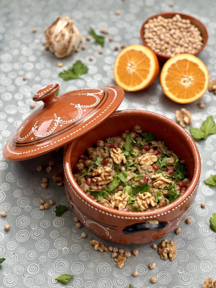 A deep clay pot with beautiful patterns full of vegan fesenjan, with the ingredients scattered around it.