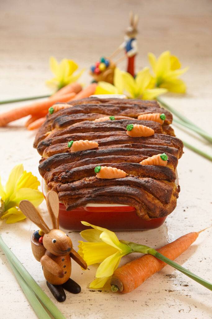 Freshly baked sweet pull apart bread decorated with small marzipan carrots in a red ceramic baking dish, with some carrots and daffodil flowers and Easter bunny figures next to it.