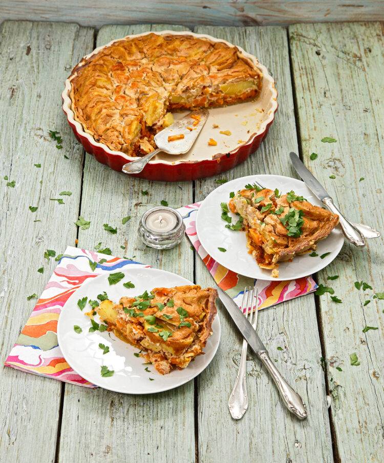 Ready-baked quiche in a tart tin that has already been cut. In front of the tart tin are two plates on which pieces of quiche have been arranged. Colorful napkins and silver cutlery.
