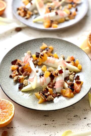 Two grey plates of Blood orange salad with Beldgian endives, nuts, mulberries, dates, and oil. Garnished with blood oranges