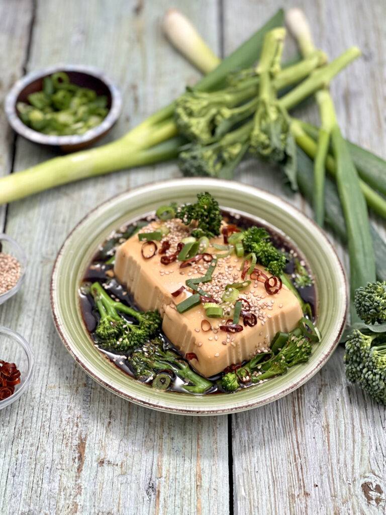 A green plate with nicely decorated cold silken tofu with fresh chives and broccolini in the background