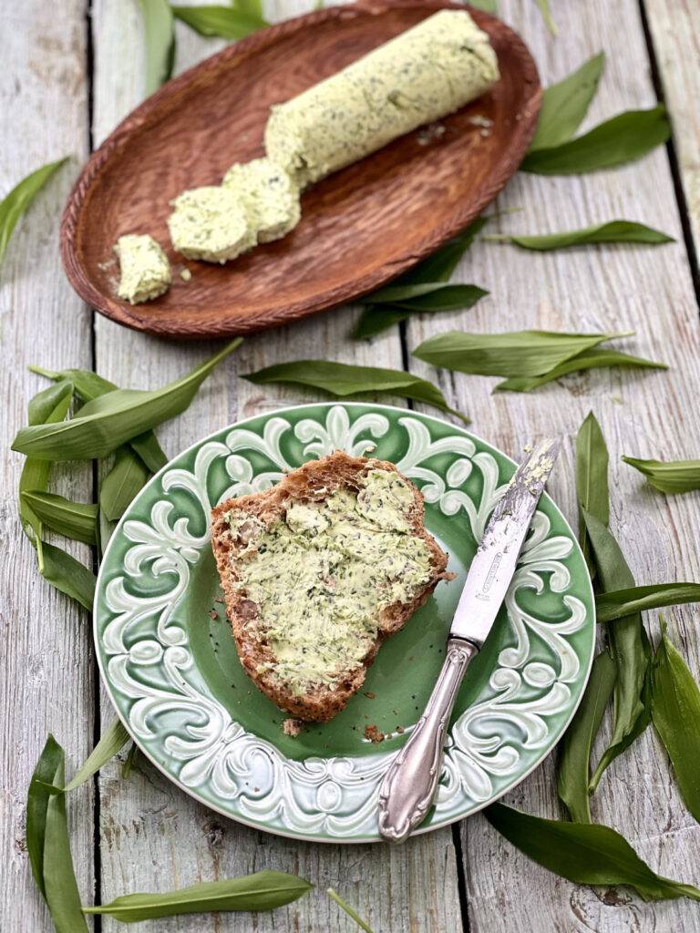 Roll of sliced wild garlic butter on a wooden tray, with a plate of bread served with wild garlic butter on a green plate