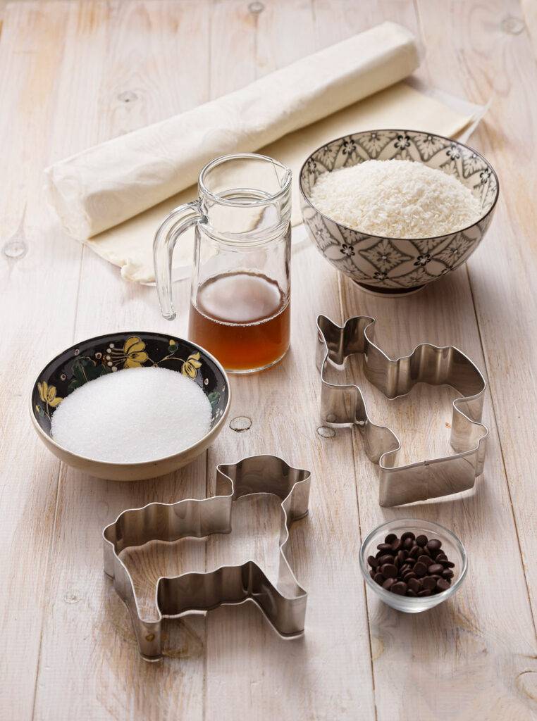 A roll of puff pastry on a light-colored wooden table. In front of it, a carafe of Aquafaber, bowls of sugar, coconut flakes, chocolate drops and two large cookie cutters.