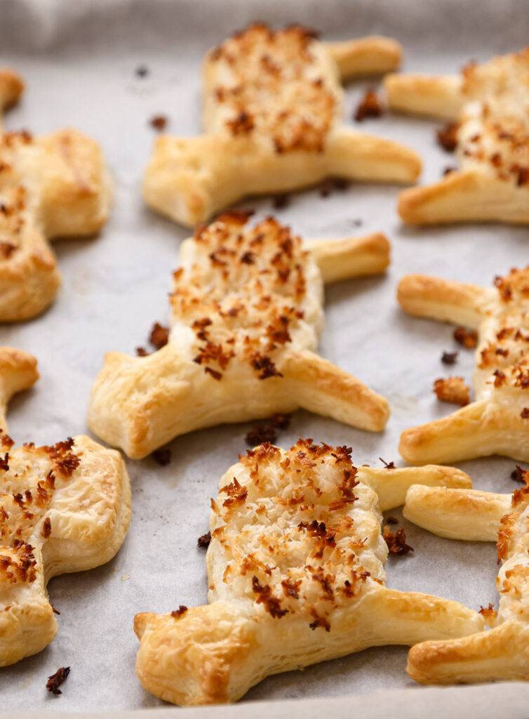 A baking tray with baking paper, lined with ready-baked puff pastry cookies in the shape of lambs and Easter bunnies, topped with grated coconut.