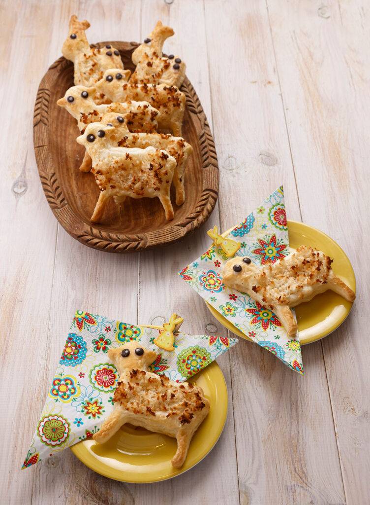 A shallow wooden bowl on a light-colored wooden table with ready-made puff pastry cookies in the shape of lambs and Easter bunnies. In front of it, two small yellow plates with napkins and lamb cookies.