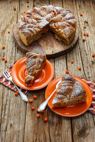 The Yeast Cake is served on a wooden board. The base consists of wooden planks on which a few sea buckthorn berries are scattered. The yeast cake is cut, pieces of cake lie on the two orange-colored plates in the foreground, next to forks and napkins.