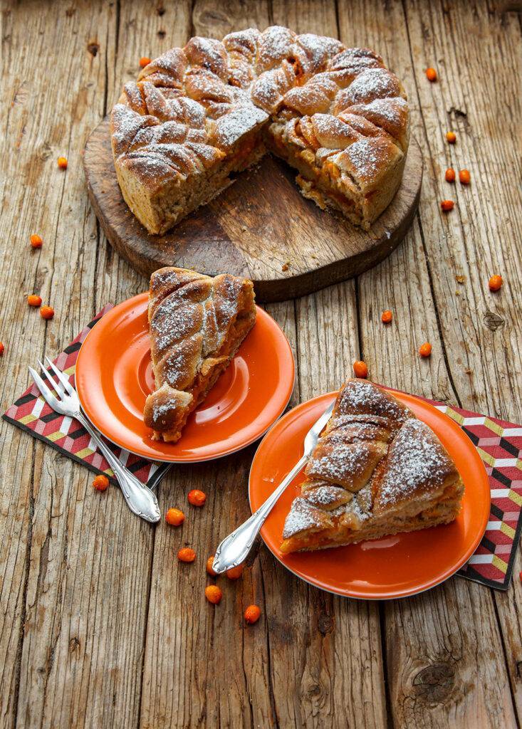 The Yeast Cake is served on a wooden board. The base consists of wooden planks on which a few sea buckthorn berries are scattered. The yeast cake is cut, pieces of cake lie on the two orange-colored plates in the foreground, next to forks and napkins.