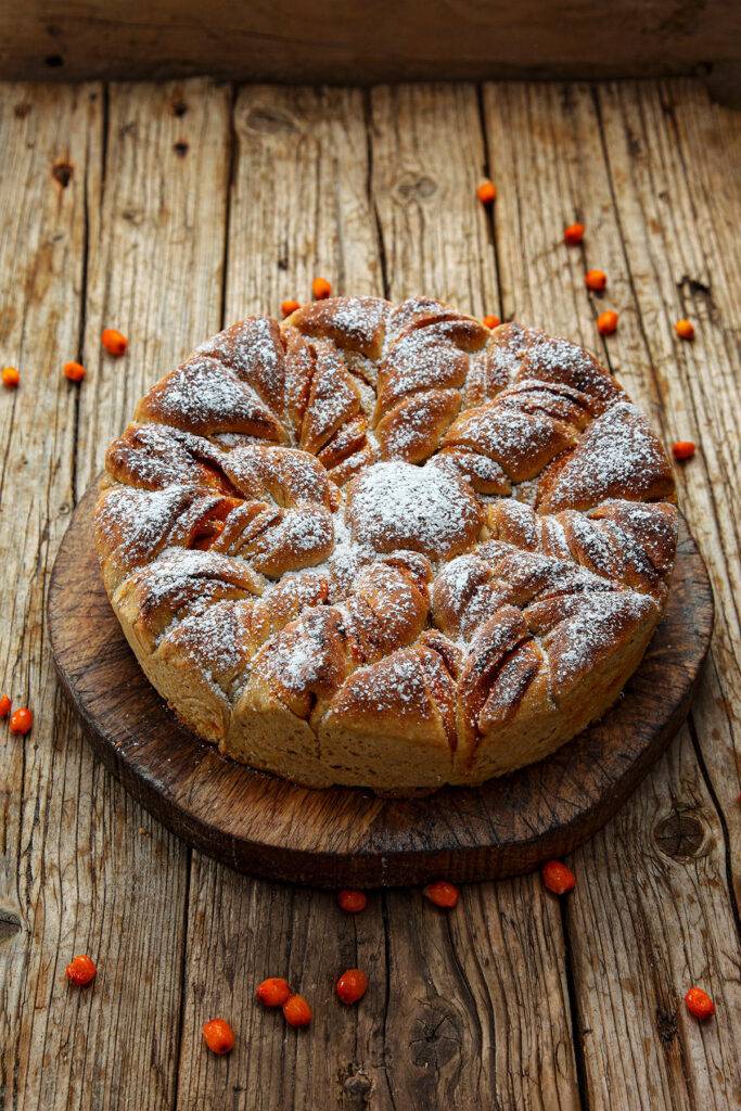 The Yeast Cake is served on a wooden board. The base consists of wooden planks on which a few sea buckthorn berries are scattered.