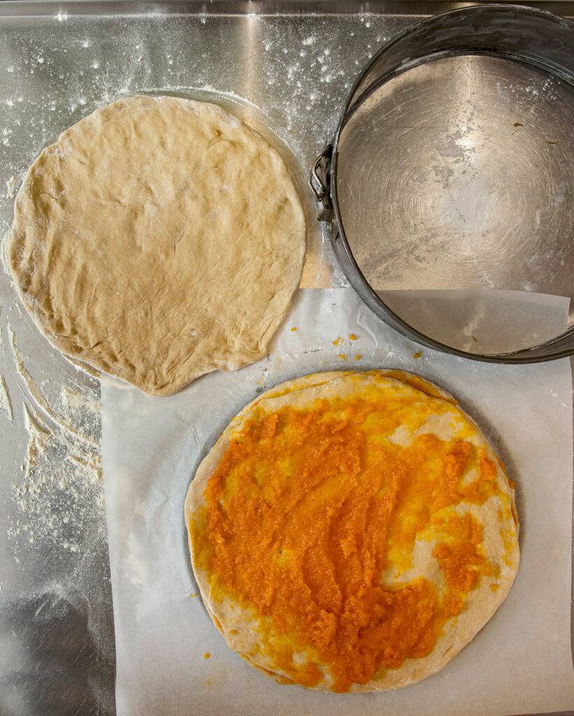 The yeast cake is layered, you can see a springform ring, yeast dough bases and leftover flour on baking paper - all on a stainless steel plate. A yeast dough base is topped with sea buckthorn puree.