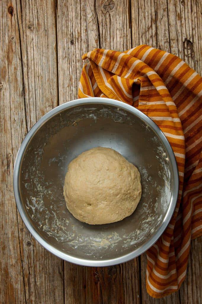 The ball of yeast dough lies in a stainless steel bowl. The base is made of wooden planks. Behind the bowl is a tea towel.