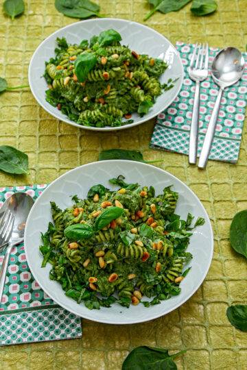 Two white plates filled with vegan pasta and spinach, topped with pine nuts and basil, sit on a green tablecloth. Napkins and cutlery are placed next to the plate. Basil leaves are scattered about.
