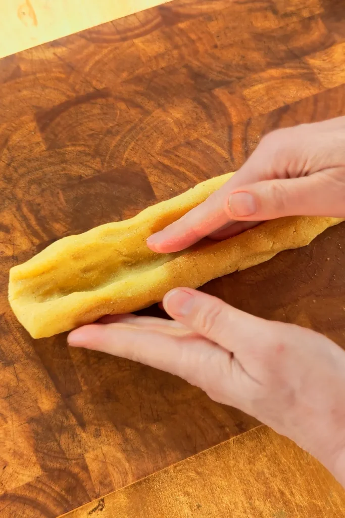 One hand prepares a trench in the dough roll. The dough roll for the Date cookies lies on a wooden board.