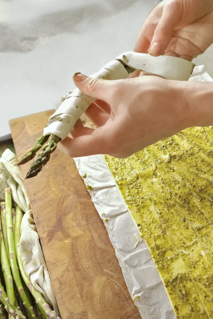 Two Hands wrapp the fresh Asparagus with the puff pastry strip. On the wooden underground lies more fresh asparagus and puff pastry strips with green pesto.