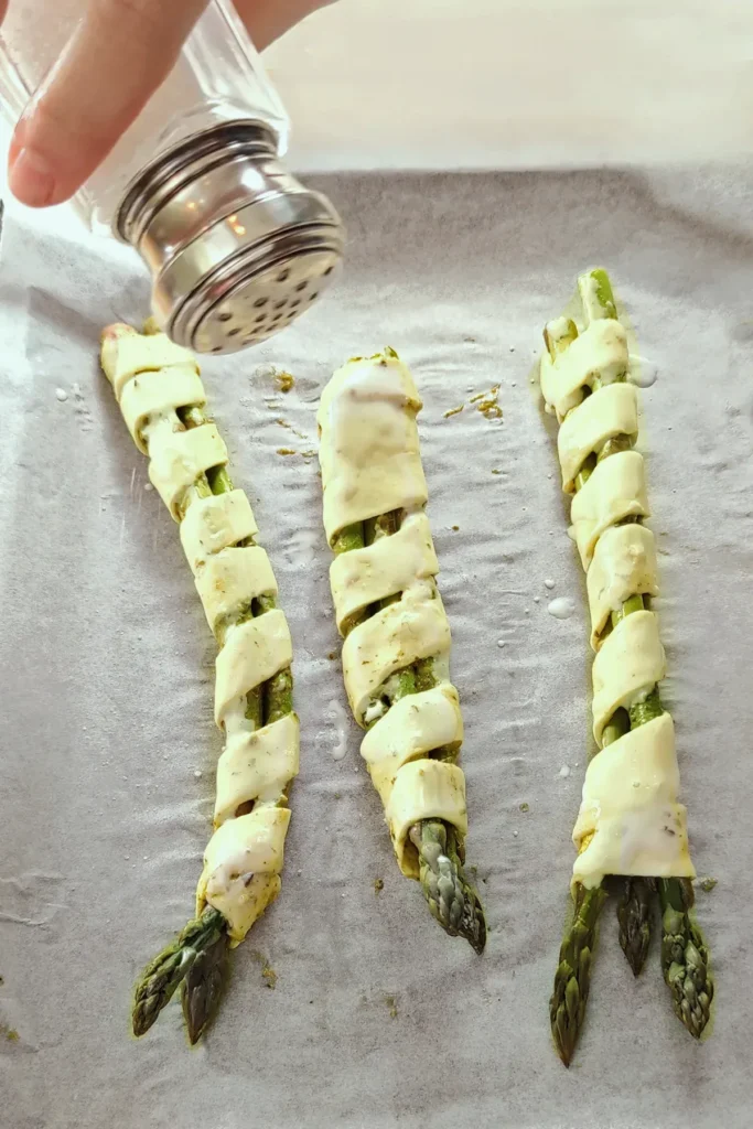 Three bundles of unbaked asparagus wrapped in puff pastry are placed on a baking tray lined with baking paper. A hand with a salt shaker salts the dough