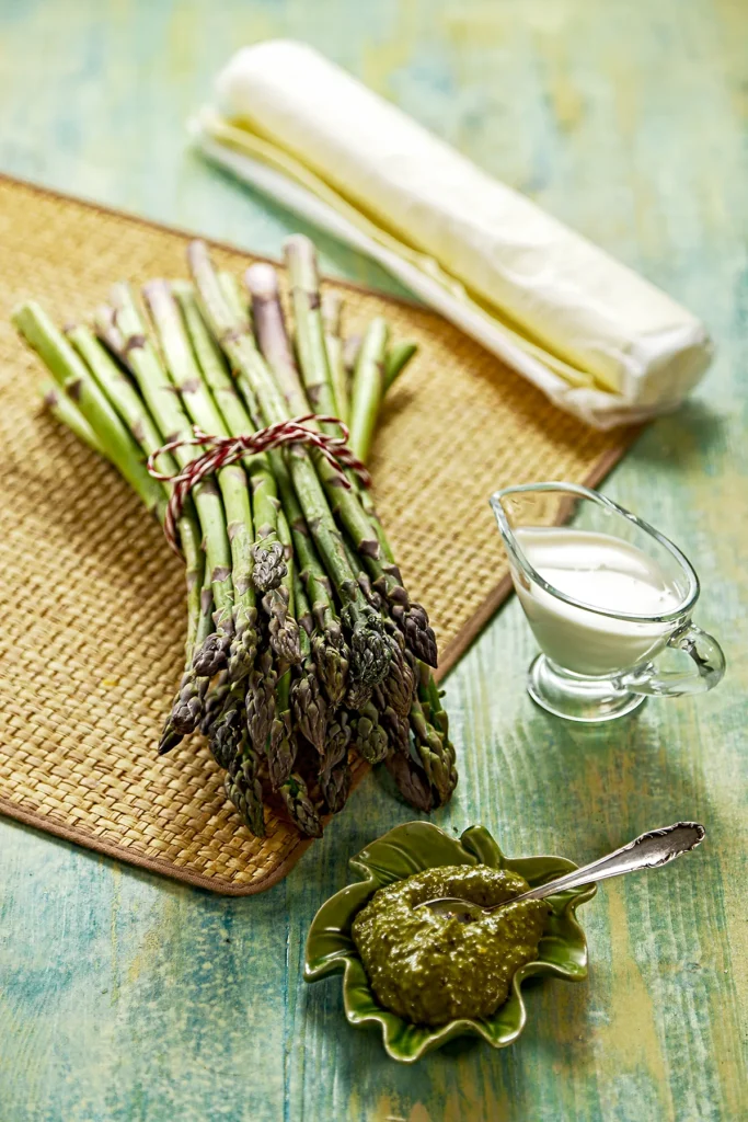 On a green wooden table with a straw mat, lies a bundle of fresh green asparagus, accompanied by a small bowl of green pesto and a jug of vegan cream. In the background, there is a roll of ready-made puff pastry.