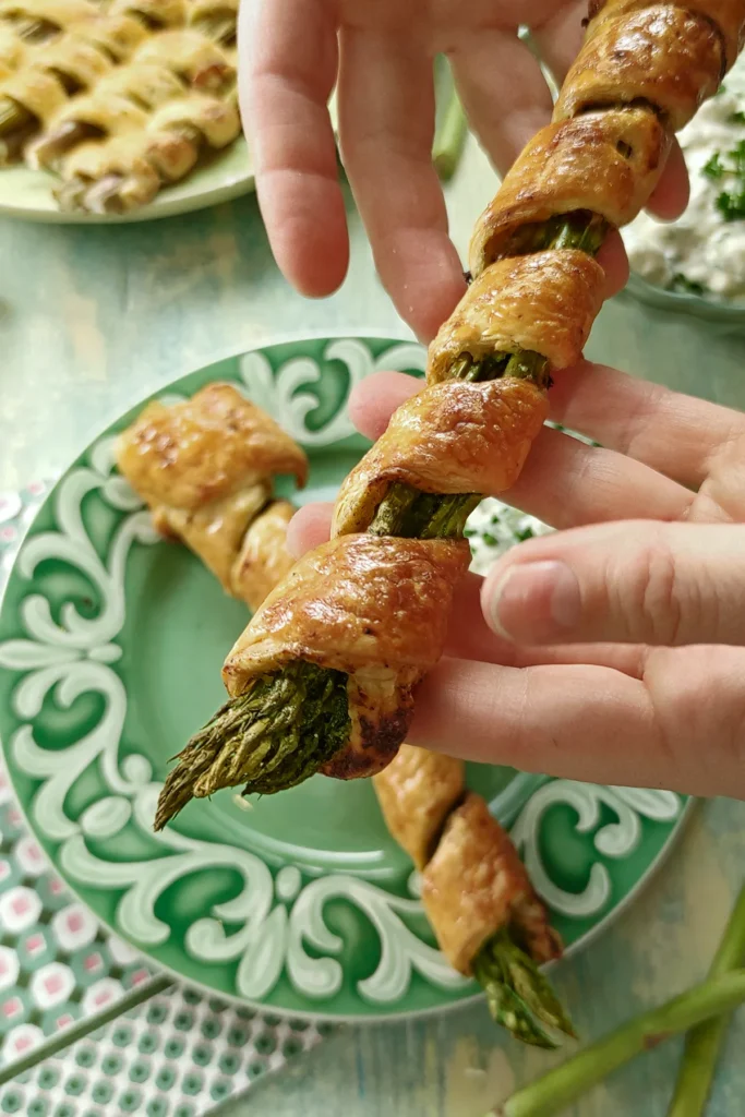 In the foreground, two hands present a baked asparagus in puff pastry. Below, a plate of baked asparagus in puff pastry. In the background, a serving plate with more baked asparagus in puff pastry and a bowl of cucumber yogurt can be seen out of focus.