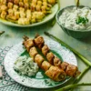 In the foreground there is a plate with two baked asparagus in puff pastry. In the background there is a serving plate with more baked asparagus in puff pastry and a bowl of cucumber yogurt can be seen out of focus