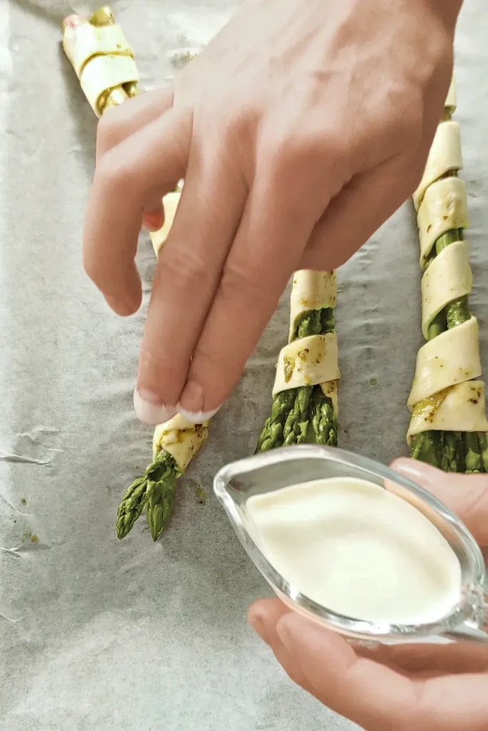 Three packets of asparagus in puff pastry are placed on baking paper, the puff pastry strip is drizzled with vegan cream.