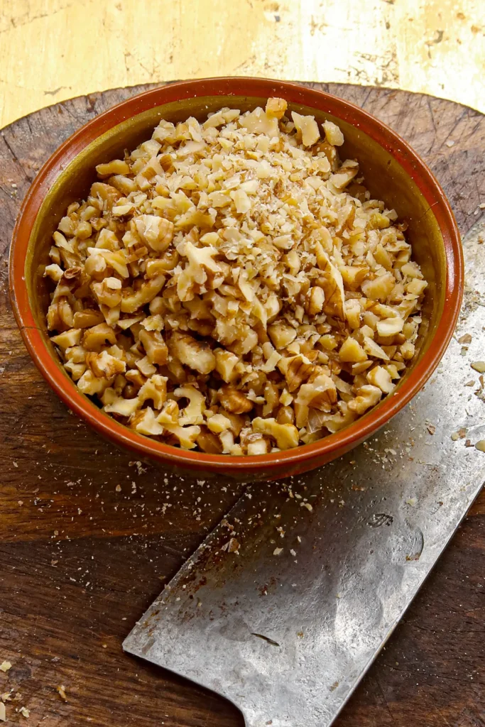 A bowl of chopped walnuts for the Date cookie filling is placed on a wooden board with a knife next to it.