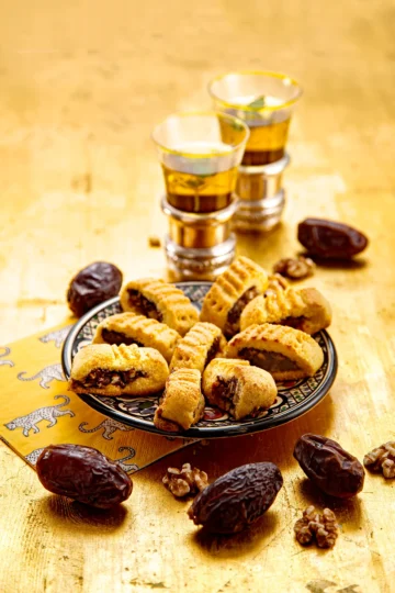 Date cookies presented on a plate, two cups of tea in the background. Dates and walnuts are scattered around the plate on a golden wooden underground.