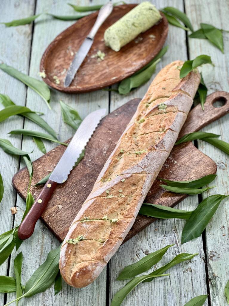A sliced block of wild garlic butter and a fresh baguette filled with wild garlic butter on a wooden underground