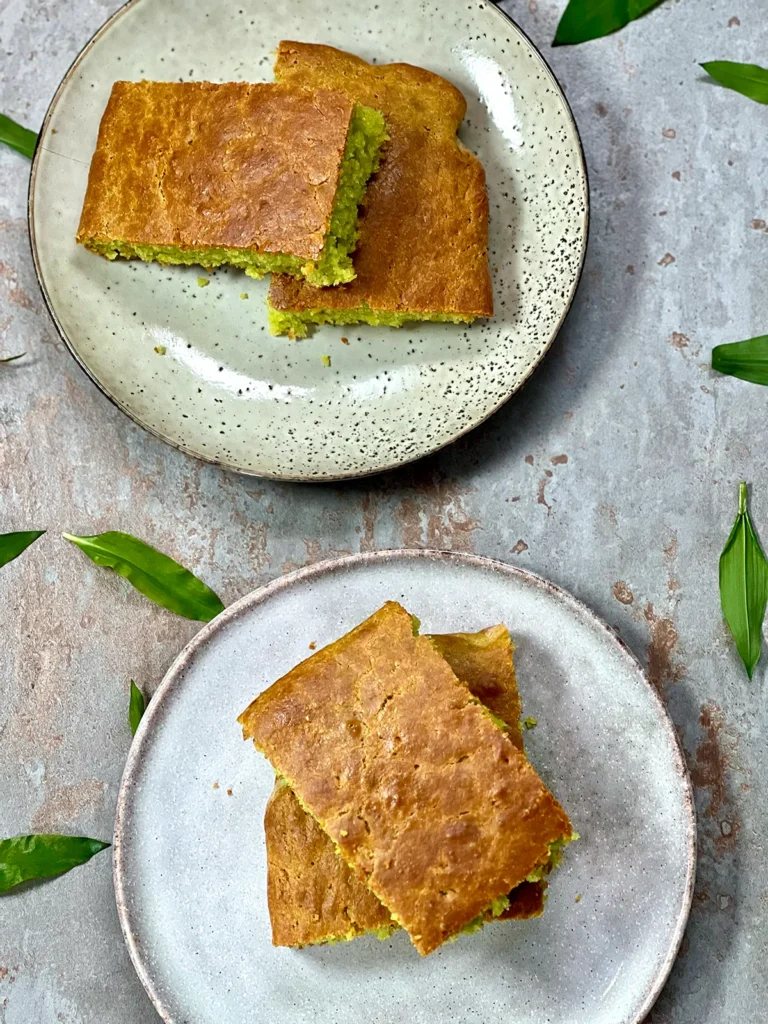 Top angle shot of four pieces of wild garlic focaccia with a delicious brown crust. Served on two plates on a dark stone plate with wild garlic leaves around them.