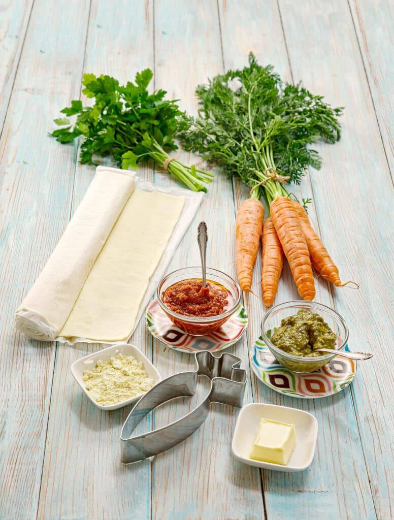 The ingredients for the Puff Pastry Carrots recipe are arranged on a wooden board: fresh carrots, parsley, puff pastry, red and green pesto, vegan butter, and egg substitute powder. Also included is a carrot-shaped cookie cutter.