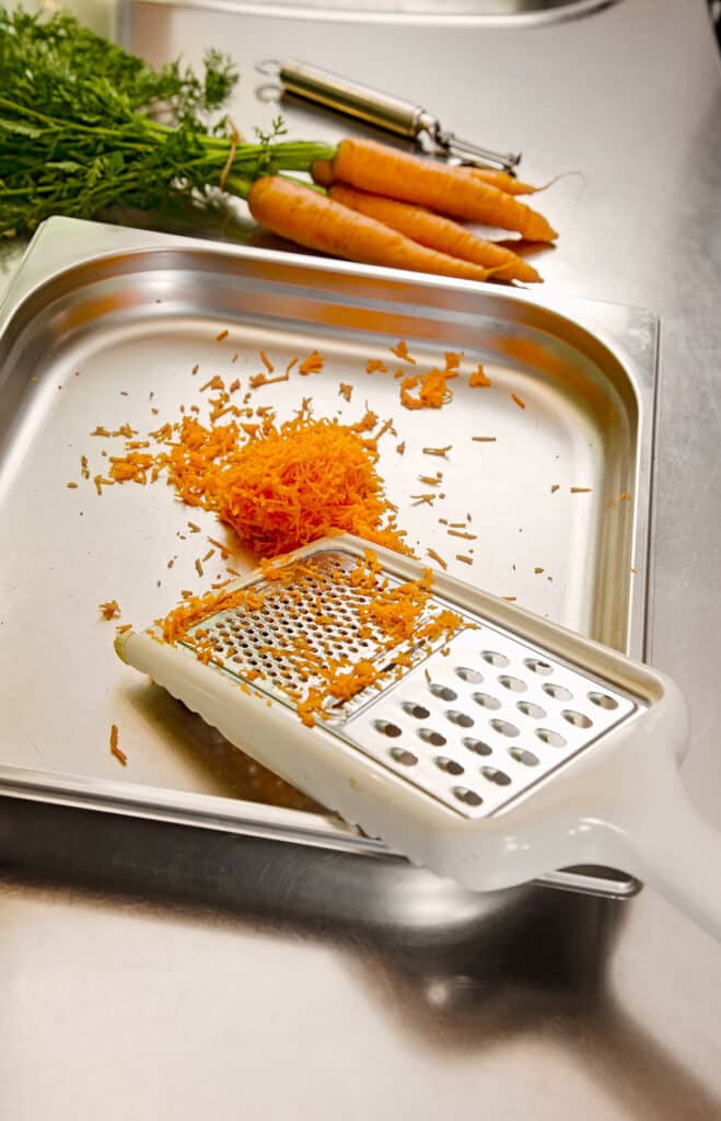 A stainless steel bowl holds a grater and freshly grated carrots. In the background, there is a bunch of fresh carrots and a peeler.