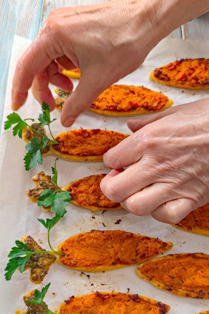 Puff pastry carrots that are ready-baked on baking paper are garnished with parsley using both hands.