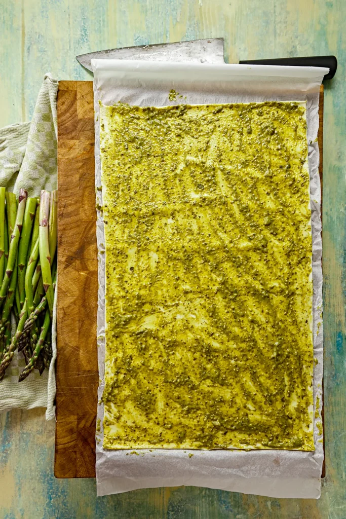 The puff pastry is rolled out on baking paper and completely covered with green pesto. Fresh asparagus is placed next to it on a towel.