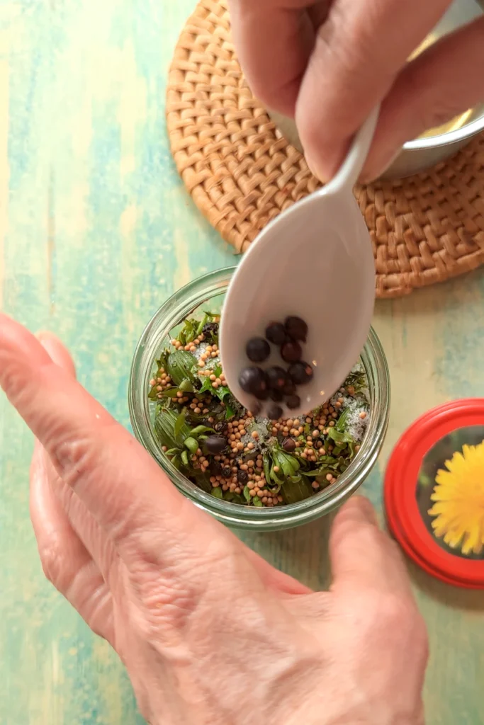 One hand holds a spoonful of spices and sprinkles them into the jar of dandelion buds.