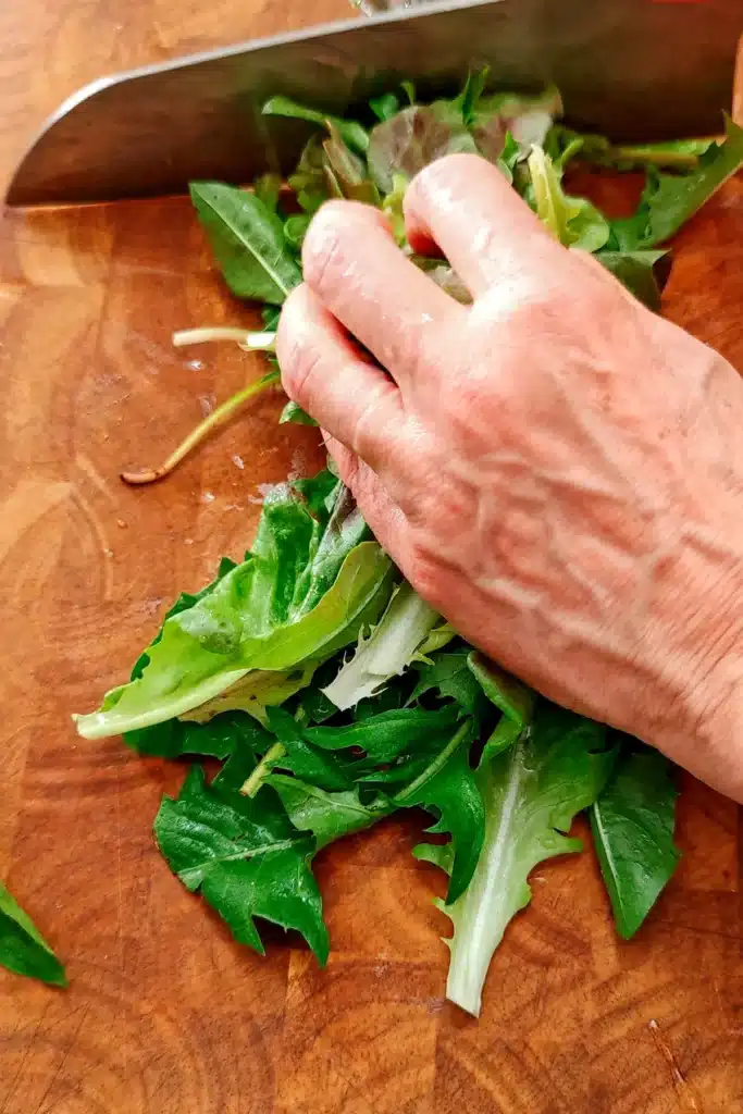 A hand holds dandelion leaves on a wooden board. A huge knife is chopping the leaves.