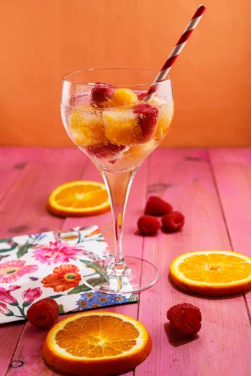 A glass of water with fruit ice cubes and a straw stands on a pink wooden base. Next to the glass are slices of oranges, raspberries and a napkin.