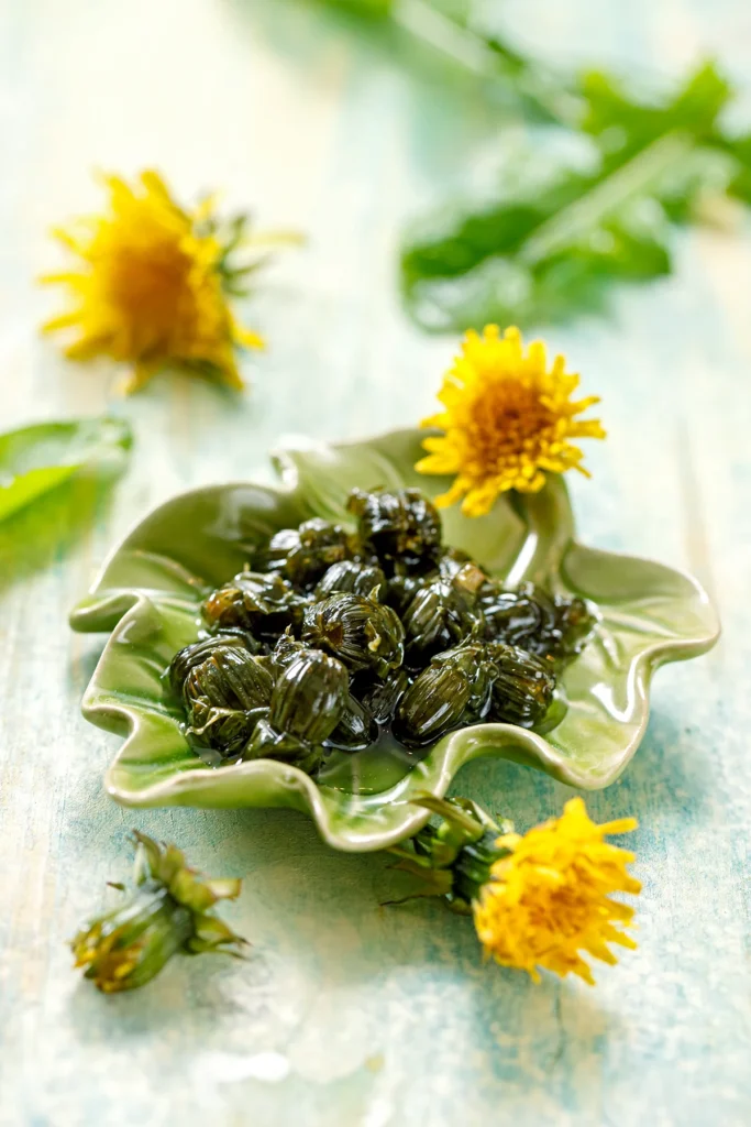 The pickled dandelion buds are arranged in a bowl. There are dandelion leaves and flowers decorated around.
