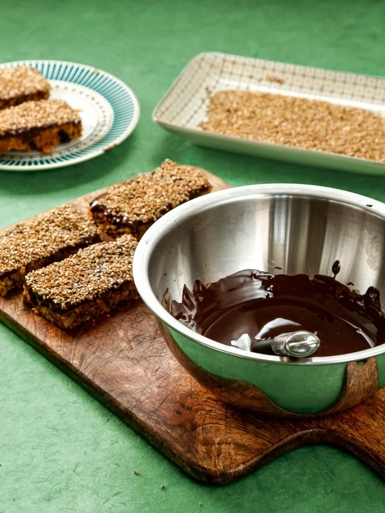 On a wooden cutting board are lying three ready cereal bars with topping. In front is a metal bowl with melted chocolate. Behind the cutting board, there is a plate with two more cereal bars and a plate with roasted sesame seeds. All are placed on green ground.
