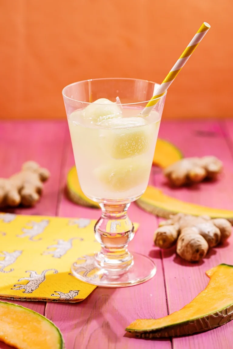 A glass with ginger melon ice cubes and a straw stands on a pink wooden base. Next to the glass is are slices of melon and ginger roots and a napkin.