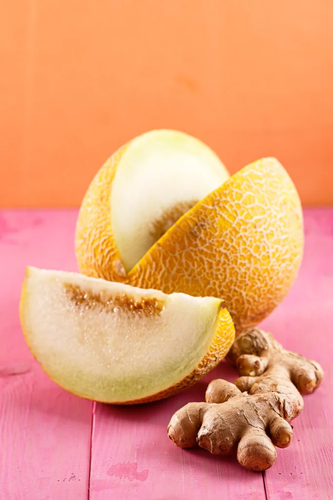 A sliced Galia melon and ginger root are lying on a pink wooden base.