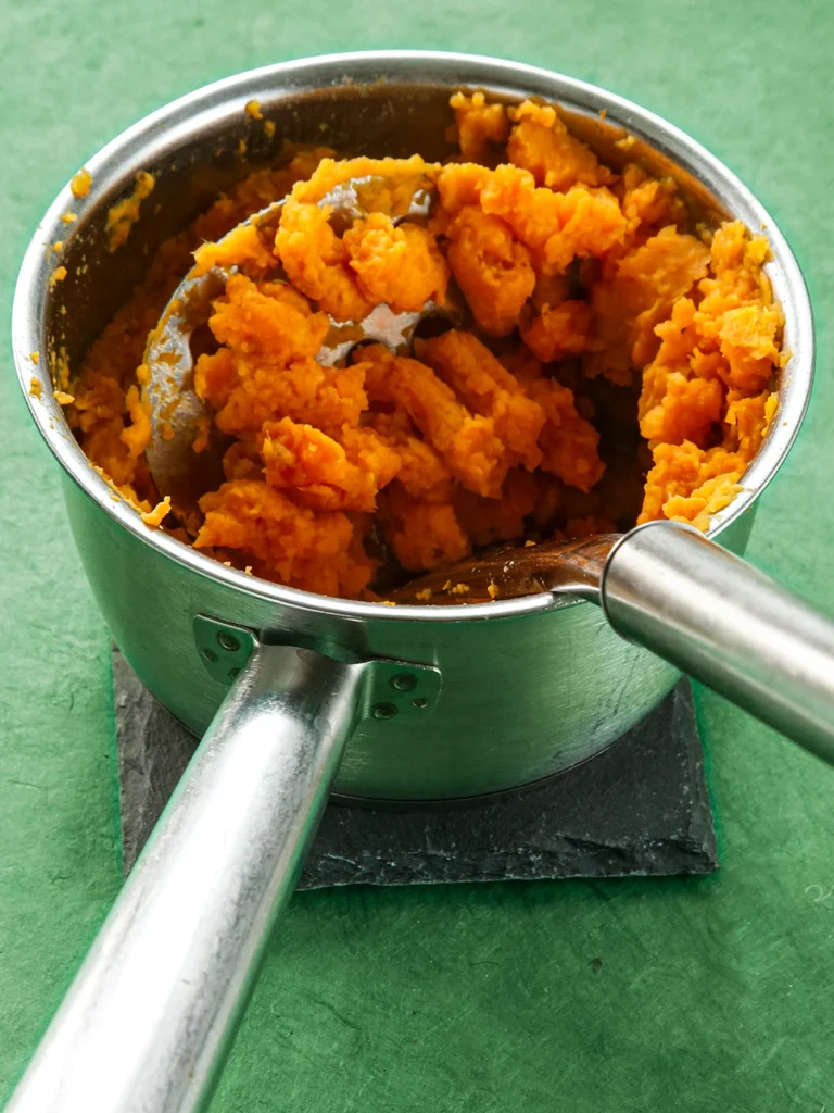 Mashed sweet potato in a pan with a potato masher on a green ground.