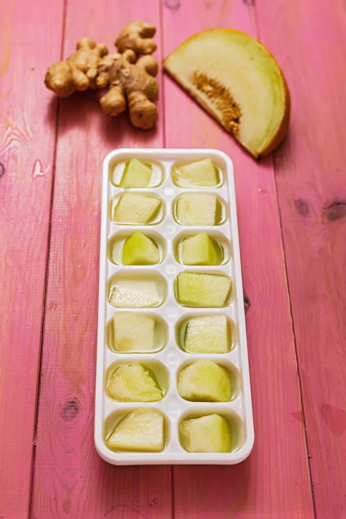 A ice cube mold with ginger juice and pieces of galia melon are standing on a pink wooden underground. Behind the ice cube mold are a ginger root and a slice of melon.