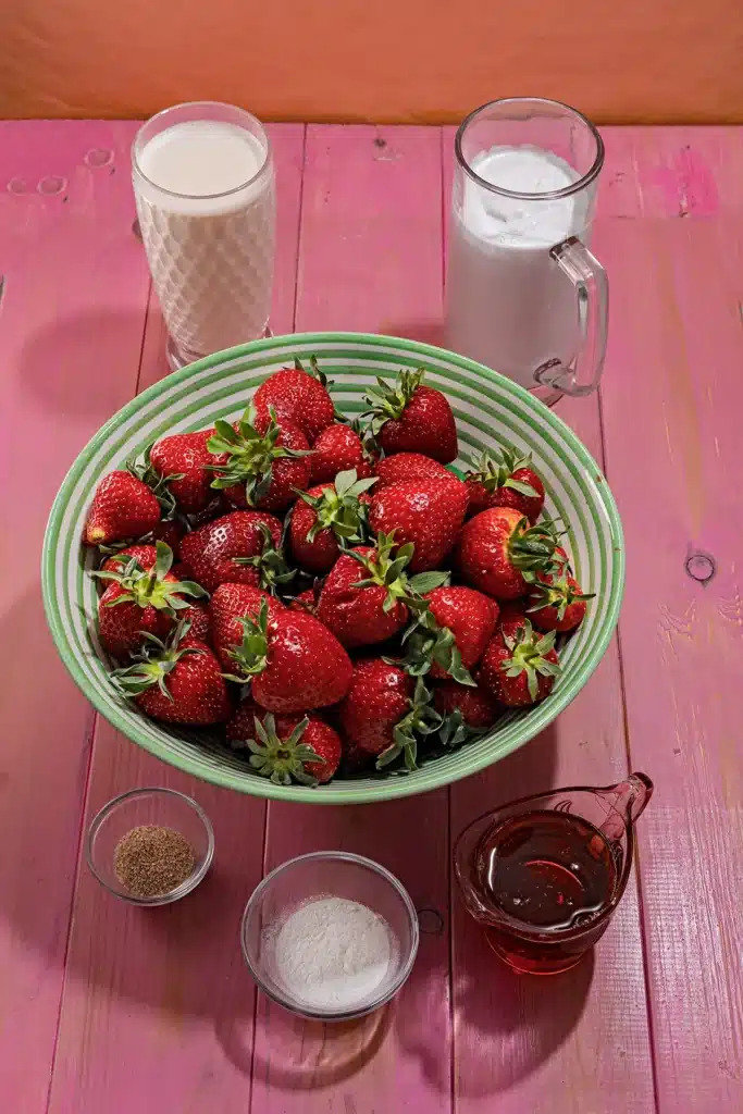 A green and white striped bowl with fresh strawberries stands in the middle on a pink wooden background. Behind the bowl are two glasses of coconut milk and oat milk. In front of them are two small bowls of vanilla sugar and agar agar, and a small jug of maple syrup.