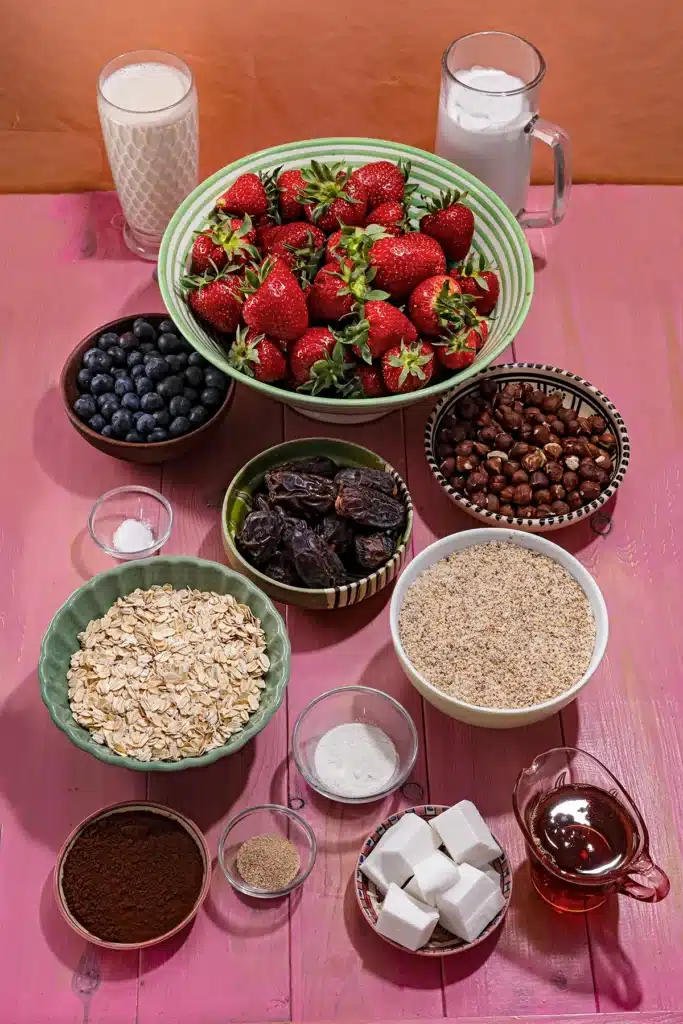 Various bowls with rolled oats, hazelnuts, ground hazelnuts, medjool dates, cocoa, pieces of palm oil, dark chocolate, fresh strawberries, two glasses with coconut milk and oat milk, tree small bowls of salt, vanilla sugar and agar agar, and a jug of maple syrup stand on a pink wooden background.